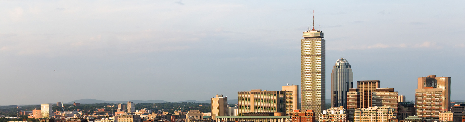 The image shows a city skyline with a prominent skyscraper in the foreground, viewed from across a body of water during what appears to be either sunrise or sunset, given the warm lighting and reflections on the water surface.