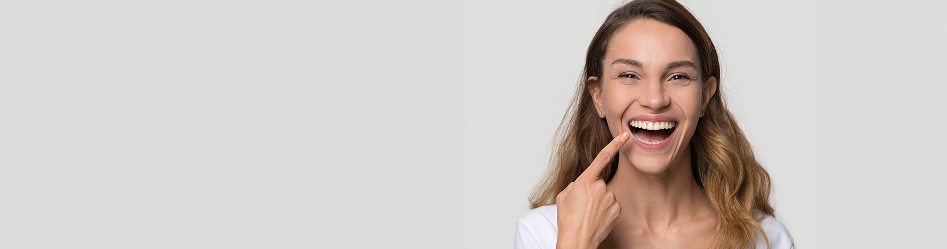 The image is a photograph of a woman with light skin, smiling at the camera. She appears to be in her late twenties or early thirties and has long hair. Her eyes are looking directly at the camera, and she is holding up her index finger near her mouth as if she s making a point or emphasizing something. The background is plain and light-colored, which suggests that this could be a stock photo used for various purposes such as advertising, personal branding, or lifestyle content.