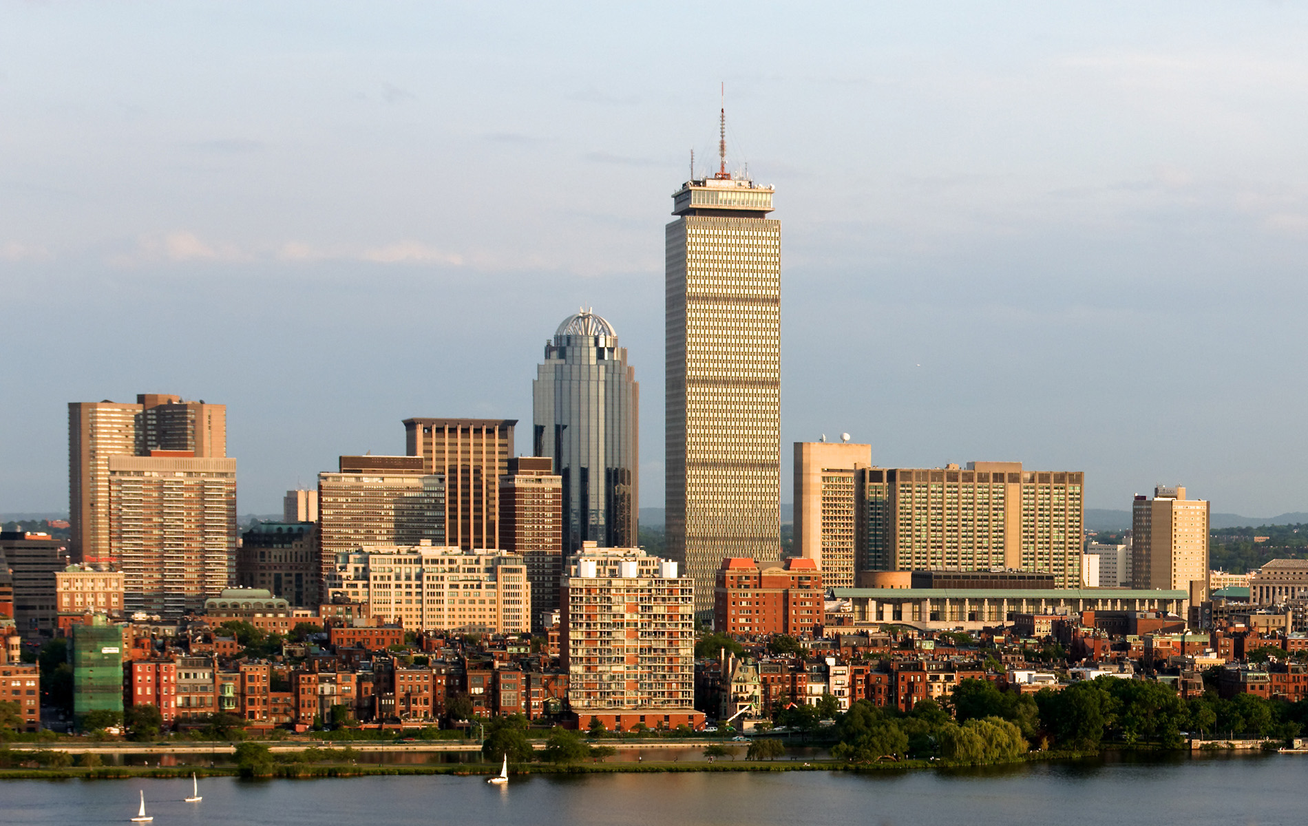 The image shows a panoramic view of a city skyline with tall buildings under a clear sky, featuring a prominent skyscraper, the Boston skyline, and a river in the foreground.