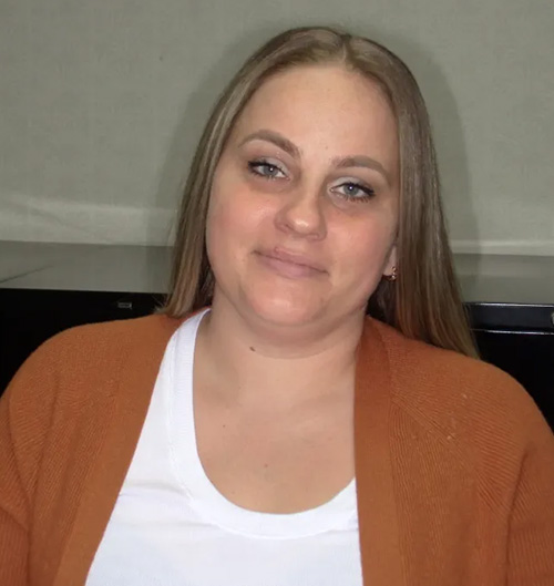 The image shows a woman with light-colored hair, wearing a brown sweater, sitting in front of a white wall.