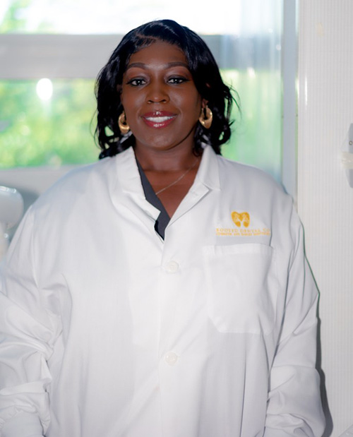 The image shows a woman wearing a white lab coat with a name tag, smiling at the camera.