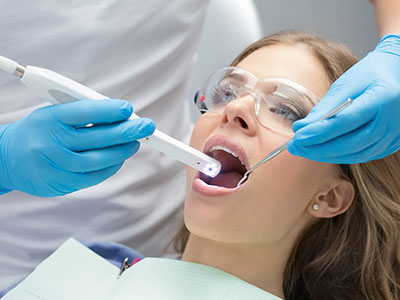 The image shows a woman sitting in a dental chair with her mouth open, receiving dental care. A dental professional is using an electronic device to examine her teeth, assisted by another person wearing protective gloves and glasses.